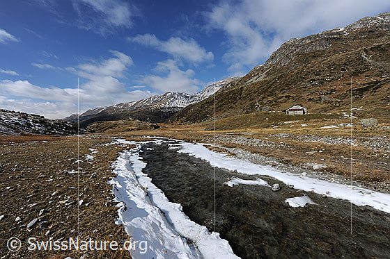 Foto: Weite Hochebene mit Bergbach. Schneereste von erstem Schnee im Herbst säumen das Bachufer. Über der herbstlichen Berglandschaft sind lockere Quellwolken zu sehen.