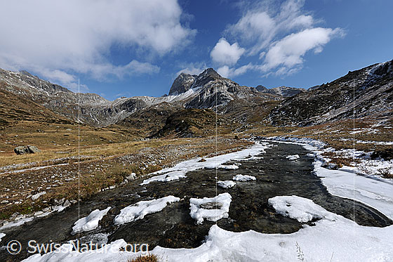 Foto: Schneereste entlang eines Bergbachs auf urtümlicher, weiter Hochebene. Über der Berglandschaft mit Ofenhorn reissen die Wolken auf.