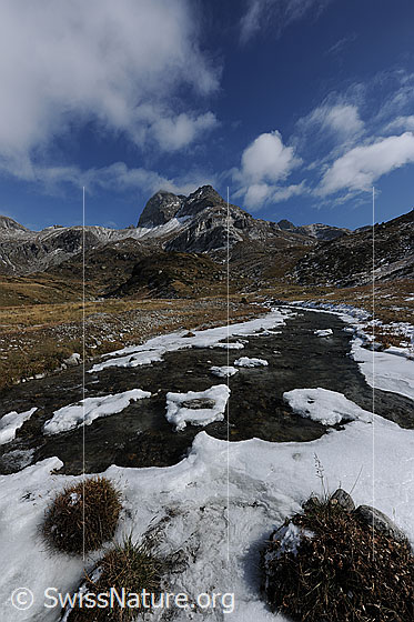 Foto: Binna mit Schneeresten am Ufer. Der Bergbach fliesst flach über die herbstliche Hochebene. Am blauen Himmel über dem Ofenhorn sind stimmungsvolle Wolkenfetzen zu sehen.