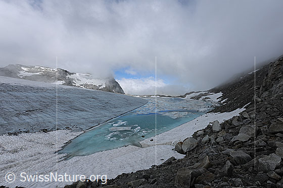 Foto: Wolkenstimmung über Gletschersee und Chüebodengletscher. Urtümliche, geheimnisvolle Gletscherlandschaft mit Wolkendecke. Auf dem türkisfarbenen See sind Eisschollen und eine schmelzende Eisdecke zu sehen. Im klaren Wasser ist Gletschereis erkennbar. Die Eisoberfläche des Gletschers ist ausgeapert und bildet eine scharfe Kante dem Seeufer entlang.