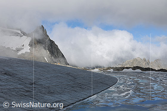 Foto: Chüebodengletscher und Gletschersee mit Wolkenstimmung. Die Wolkendecke hat sich angehoben und gibt den Blick auf den ausgeaperten Gletscher, den See mit den Eisschollen und den Ausläufer des Poncione di Manio frei. Eine ausgeprägte Kante aus Eis formt das Ufer des Bergsees. In der Eisfläche sind die Jahrringe deutlich zu sehen.