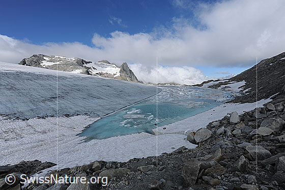 Foto: Gletscherlandschaft mit Wolkenstimmung. Urtümliche Berglandschaft bestehend aus Blockgelände, Gletschersee im Gletschereis und Eisschollen, welche sich von der aufgebrochenen Eisdecke gelöst haben. Das Wasser hat die Farbe türkis. Durch das Abschmelzen des Chüebodengletschers vergrössert sich der See laufend (Gletscherschwund, Klimawandel). Mittlerweile misst der See in der Breit ca. 100m. Im Jahr 2008 waren erst zwei sehr kleine Seen vorhanden.