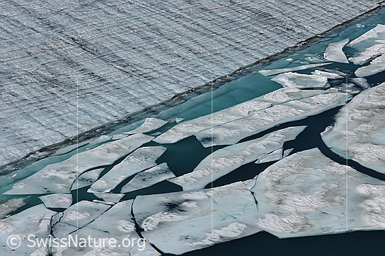 Foto: Eisschollen auf Gletschersee. Das Wasser am Gletscherrand hat die Farbe türkis. Der Eisrand bildet eine Linie und die Schollen sind in unterschiedliche Formen zerbrochen. Auf der Eisfläche des Gletschers sind die Jahrringe zu sehen.