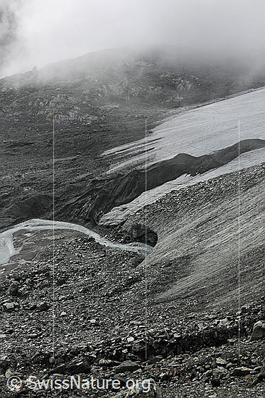 Foto: Gletschertor und Nebelstimmung. Das Gletschereis über dem Tor ist mit Geröll bedeckt. Die Nebelreste hüllen die Gletscherlandschaft mit Gletscherbach in eine geheimnisvolle Stimmung.