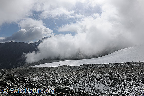 Foto: Wolkenstimmung über Gletscherlandschaft des Schwarzberggletschers. Blick über das mit Geröll bedeckte Gletschereis am Gletscherrand auf den Gletscher. Die Restbewölkung löst sich stimmungsvoll auf.