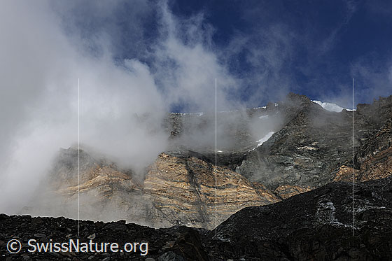 Foto: Wolkenfetzen und Felswand. Stimmungsvoll aufreissende Restwolken an den Felsen des Schwarzberg und den Felswänden und Graten von Strahlhorn und Fluchthorn. Die Seitenmoräne und das mit Geröll bedeckte Gletschereis liegen noch im Schatten.