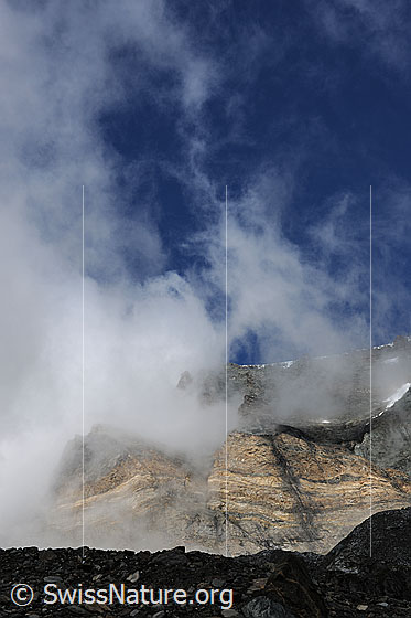Foto: Felswand und Wolkenfetzen. Stimmungsvoll aufreissende Restwolken an den Felsen von Schwarzberg und Strahlhorn. Die Seitenmoräne und das mit Geröll bedeckte Gletschereis liegen noch im Schatten.