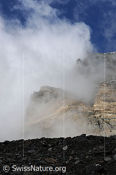 Foto: Wolkenstimmung am Strahlhorn. Blick vom Wandfuss in die eindrückliche Felswand mit Graten und Felsköpfen, welche durch die aufreissenden Wolken sichtbar werden. Vordergrund: Geröllwall der Seitenmoräne des Schwarzberggletschers.