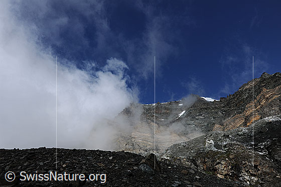 Foto: Strahlhorn und Fluchthorn mit Wolkenstimmung. Am Fuss der eindrücklichen Felswand sind Moräne und Gletschereis des Schwarzberggletschers zu sehen. An den Felsen und Graten ziehen Wolkenfetzen vorüber.