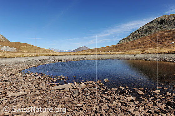 Foto: Undre Rossusee im Herbst. Der Bergsee ist fast ausgetrocknet. Steine und Alpweiden umgeben den blauen See auf der weiten Hochebene.