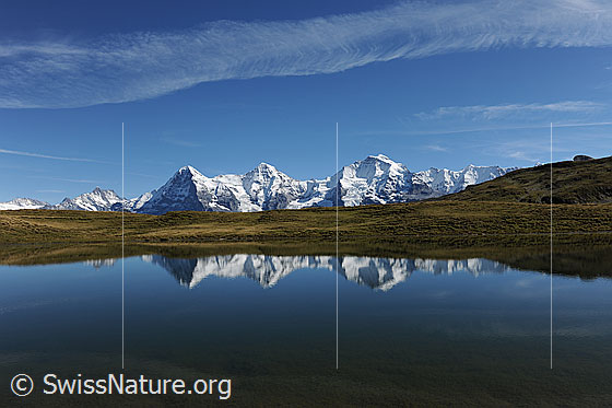 Foto: Spiegelbild des Dreigestirns in Bergsee. Ein Wolkenband aus Schleierwolken dekoriert den blauen Himmel.