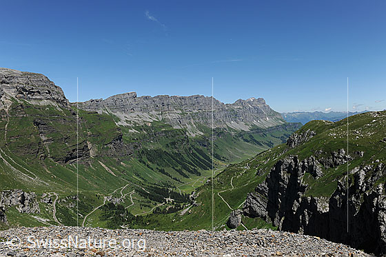 Foto: Urnerboden, Läckistock, Jegerstock und Ortstock.
Tiefblick auf die Passstrasse zum Klausenpass und auf den Urnerboden im Taleinschnitt. Die Berghänge und Alpweiden sind grün. Im Hintergrund ist das Felsmassiv mit Läckistock, Jegerstöck und Ortstock zu sehen.
