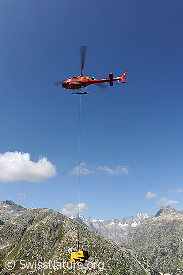 Foto: Helikopter im Einsatz für die Sanierung eines Bergwegs. 
Der am Seil befestigte Kompressor wird umplaziert.