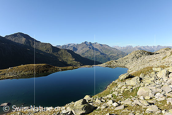 Foto: Blauer See in Berglandschaft mit Gemsstock. Auf der Alpweide um den Bergsee liegen Felsblöcke.