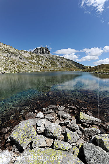 Foto: Am Ufer des Lutersee. Blick über die Steine am Ufer und die Spiegelung in der ruhigen Wasserfläche des Bergsees zum Schneehüenerstock.