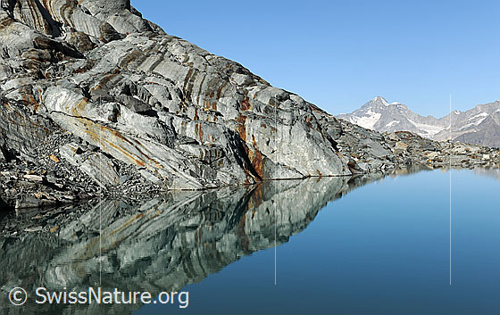 Foto: Spiegelbild im Alphubelsee. Im ruhigen, blauen Wasser des Bergsees spiegelt sich die interessant geformte und geschliffene Felsschichtung. Dabei entstehen gleichmässige Muster.