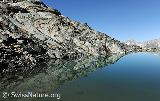 Foto: Spiegelung im Alphubelsee. Im ruhigen, blauen Wasser des Bergsees spiegelt sich die interessant geformte und geschliffene Felsschichtung. Dabei entstehen gleichmässige Muster.