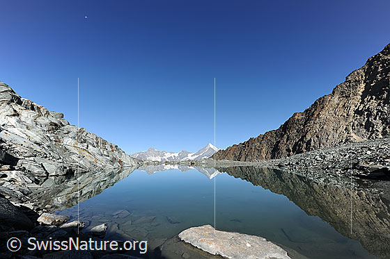 Foto: Am Ufer eines Bergsees. Blick über die spiegelnde Wasserfläche des Alphubelsees zu Zinalrothorn und Weisshorn. Die Spiegelung der Berglandschaft mit Gletscherschliff und Felsgrat lassen interessante Formen und Muster entstehen.