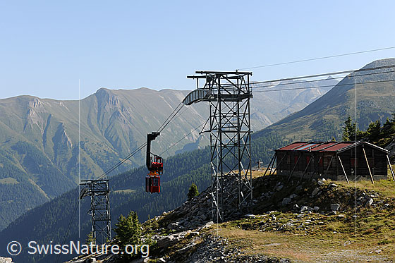Foto: Werkbahn (Luftseilbahn) unterwegs zwischen zwei Masten. An der Gratkante befindet sich eine Baracke.