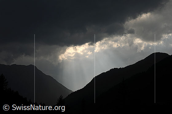 Foto: Dramatische Abendstimmung mit Regenwolken über dem Binntal. Sonnenstrahlen scheinen durch eine Lücke in der dunklen Wolkendecke.