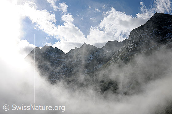 Foto: Morgenstimmung mit Nebelschwaden in Berglandschaft. Über dem mit wenig Schnee überzuckerten Felsgrat sind Quellwolken zu sehen.