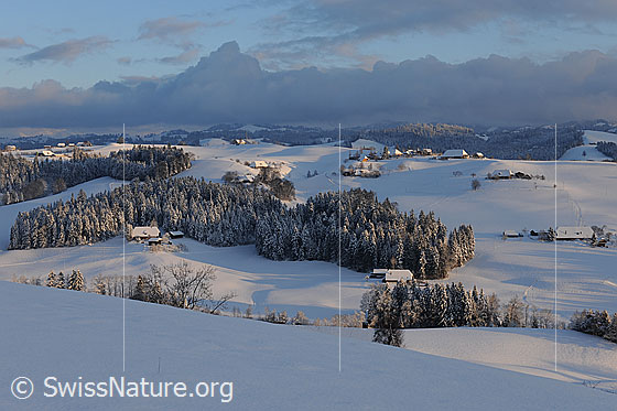Foto: Abendstimmung in verschneiter Hügellandschaft. Die Emmentaler Hügel, Wälder, Weiler und Bauernhöfe sind frisch verschneit und im letzten Abendlicht zu sehen. Im Hintergrund zieht eine Wetterfront auf.