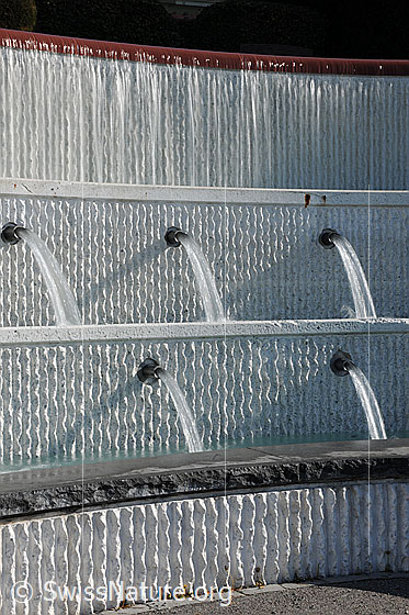 Foto: Brunnen (Langzeitbelichtung). Das Wasser fliesst als Wasserfall über die oberste Etage eines mehrstufigen Brunnens. Über mehrere Wasserstrahlen fliesst das Wasser anschliessend in die tiefer gelegenen Brunnentröge.