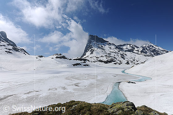 Foto: Naturlandschaft mit gefrorenem Bergsee. Die Eisdecke dem Ufer entlang beginnt aufzutauen und es hat sich ein schmaler Streifen mit hellbau gefärbtem Wasser gebildet. In der Umgebung liegt Schnee und am blauen Himmel über den Bergen sind Quellwolken zu sehen.