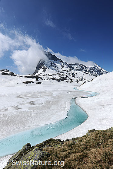 Foto: Hellblaues Wasser in eisbedecktem Bergsee. Die Eisdecke dem Ufer entlang beginnt aufzutauen. Es hat sich ein schmaler, hellbau gefärbter Wasserstreifen gebildet. In der Umgebung liegt Schnee und am blauen Himmel sind Quellwolken zu sehen.
