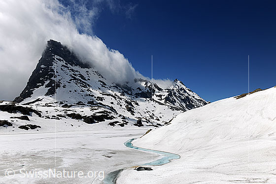 Foto: Wolkenstimmung in Berglandschaft mit zugefrorenem See. Die Eisdecke ist am Rand aufgetaut und es ist ein Streifen mit hellblauem Wasser zu sehen. Die Umgebung ist schneebedeckt.