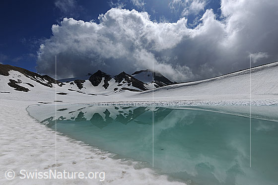 Foto: Wolkenstimmung über türkisfarbenem Bergsee. Im Wasser spiegeln sich die Berge. Über der Naturlandschaft türmen sich eindrückliche Quellwolken auf.