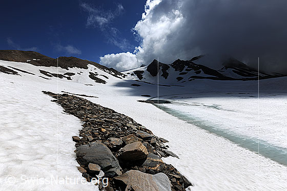 Foto: Am Ufer eines auftauenden Bergsees mit dramatischer Wolkenstimmung über der unberührten Naturlandschaft. Ein hellblauer Wasserstreifen säumt das Ufer des schneebedeckten Sees. Im Vordergrund sind kleinere Felsblöcke zu sehen.