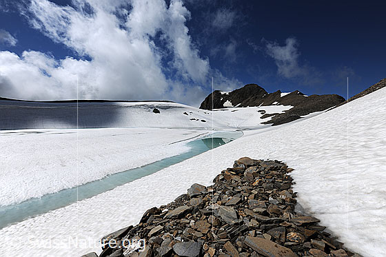 Foto: Hellblauer Wasserstreifen an auftauendem Bergsee. Am Ufer liegt Geröll und Schnee. Über der unberührten Naturlandschaft sind Quellwolken zu sehen.