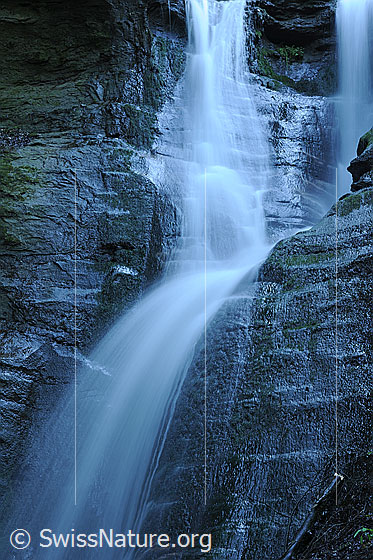Foto: Langzeitbelichtung eines Wasserfalls. Die feinen Wasserstrahlen fallen über nasse Felsen mit Strukturen.