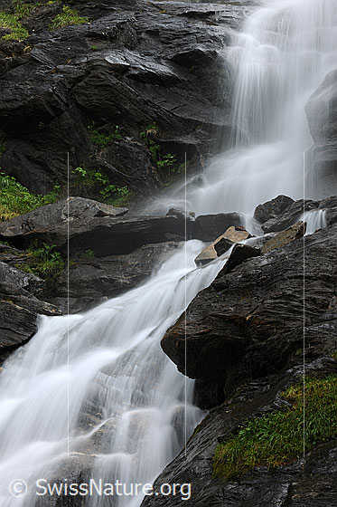 Foto: Felsen mit Wasserfall (Langzeitbelichtung). Das Wasser fällt in hellen Wasserstrahlen über die dunklen Felsen.