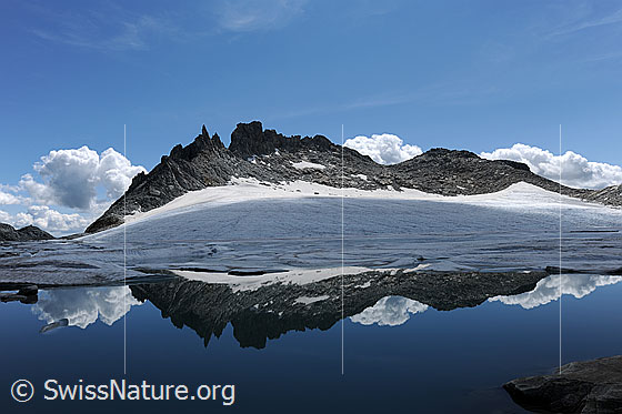 Foto: Spiegelung in Gletschersee. Ein Bergmassiv mit Gletscher im Licht und Schatten spiegelt sich in der ruhigen, blauen Wasserfläche. Im Horizont entstehen Quellwolken.