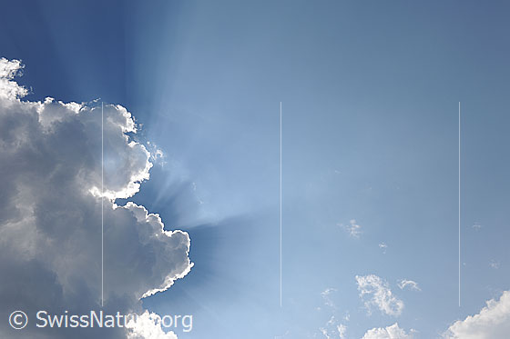 Foto: Wolkenstimmung und Lichtstrahlen am blauen Himmel. Eine dunkle Quellwolke hat sich vor die Sonne geschoben und lässt nur die Sonnenstrahlen durchscheinen. Der Rand der Wolke ist hell erleuchtet.