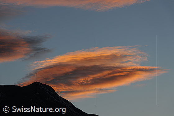 Foto: Abendstimmung mit gefärbten Linsenwolken über der Silhouette eines Berggipfels.