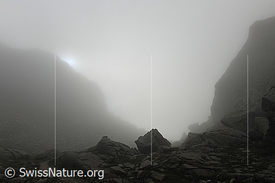 Foto: Mystische Nebelstimmung am Albrunpass. Die Sonne bricht knapp durch den Nebel und lässt die Berglandschaft als Silhouette erkennen.