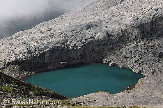 Foto: Lac de Ténéhet mit türkisfarbenem Wasser. Dahinter ein heller Felsriegel und Quellbewölkung.