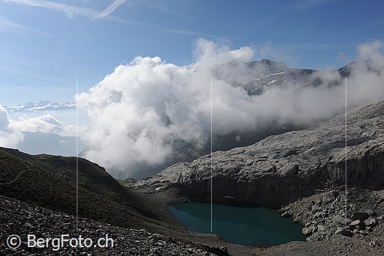 Foto: Lac de Ténéhet mit Wolkenstimmung. Ein heller Felsriegel begrenzt die kleine Mulde, in welcher der Bergsee eingebettet liegt.