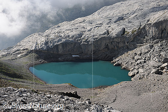 Foto: Türkis farbenes Wasser des Lac de Ténéhet. Dahinter ein heller Felsriegel und Quellbewölkung.