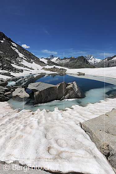 Foto: Felsblöcke in von Schnee umgebenem Bergsee.