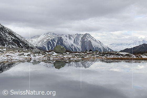 Foto: Spiegelung einer leicht verschneiten Berglandschaft. Bedeckter Himmel.