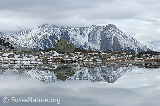 Foto: Spiegelung einer leicht verschneiten Berglandschaft in Bergsee. Bedeckter Himmel. Gemäldehaft.