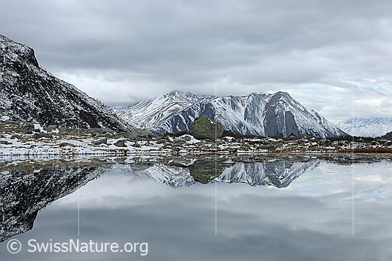 Foto: Spiegelung einer leicht verschneiten Naturlandschaft in Bergsee. Bedeckter Himmel. Gemäldehaft.