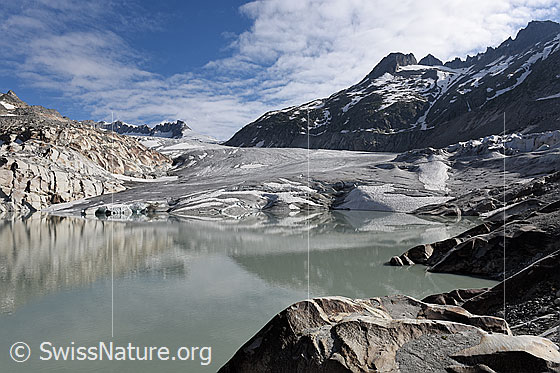 Foto: Interessante Formen an der Gletscherzunge des Rhonegletschers entstanden durch Spiegelung im Gletschersee. Die abgeflachte Zunge mündet direkt in den See. Das felsige Ufer voller Muster und Strukturen, die Schleierwolken am blauen Himmel und die schwache Spiegelung verleihen der Getscherlandschaft eine besondere Stimmung.


