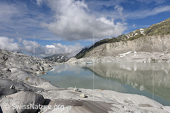 Foto: Spiegelung am Gletschersee mit stimmungsvollem Wolkenhimmel. Die Seitenmoräne des Rhonegletschers spiegelt sich im ruhigen Wasser des Rottensees. Im Vordergrund sind vom Gletscher geschliffene Felsen (Gletscherschliff aus Aaregranit) zu sehen.

